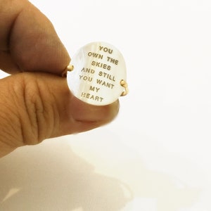 Pu&ograve; includere: Un anello d'oro con una pietra ovale bianca. La pietra &egrave; incisa con le parole "You own the skies and still you want my heart".