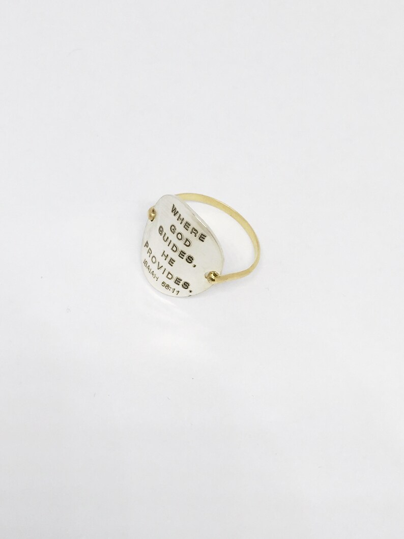 Puede incluir: Un anillo de plata con una banda de oro y un dije ovalado de plata que dice "Where God Guides, He Provides. Isaiah 58:11"