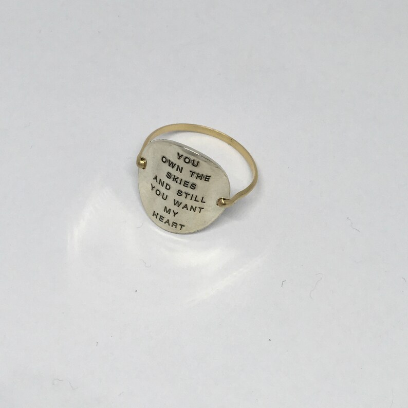 Pu&ograve; includere: Un anello d'argento con una fascia dorata e un ciondolo rotondo d'argento che dice "You own the skies and still you want my heart".