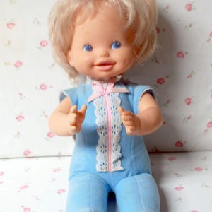 May include: A vintage baby doll with blonde hair and blue eyes. The doll wears a light blue jumpsuit with a white lace and pink zipper detail. The doll's arms are slightly raised, and it has a sweet expression.