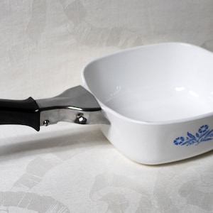 May include: White square casserole dish with a blue floral design. The dish has a metal handle with a black plastic grip. The handle is attached to the side of the dish. A classic kitchenware item.