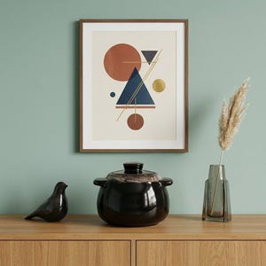May include: A framed wall art print featuring geometric shapes including circles and triangles in terracotta, navy blue, and gold on a cream background, displayed with a black ceramic lidded pot, a bird figurine, and a glass vase.