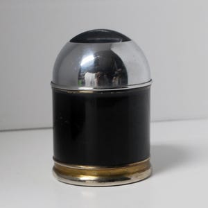 May include: A cylindrical container with a shiny chrome top and a black body. The base has a gold-colored band. The container is likely a salt or pepper shaker.