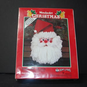 May include: A WonderArt Christmas craft kit featuring a Santa Claus design. The kit includes a red and white Santa Claus face with a red hat and white beard. The packaging is red with green and gold accents and text that reads "No Knitting. No Crocheting. Easy To Make!"