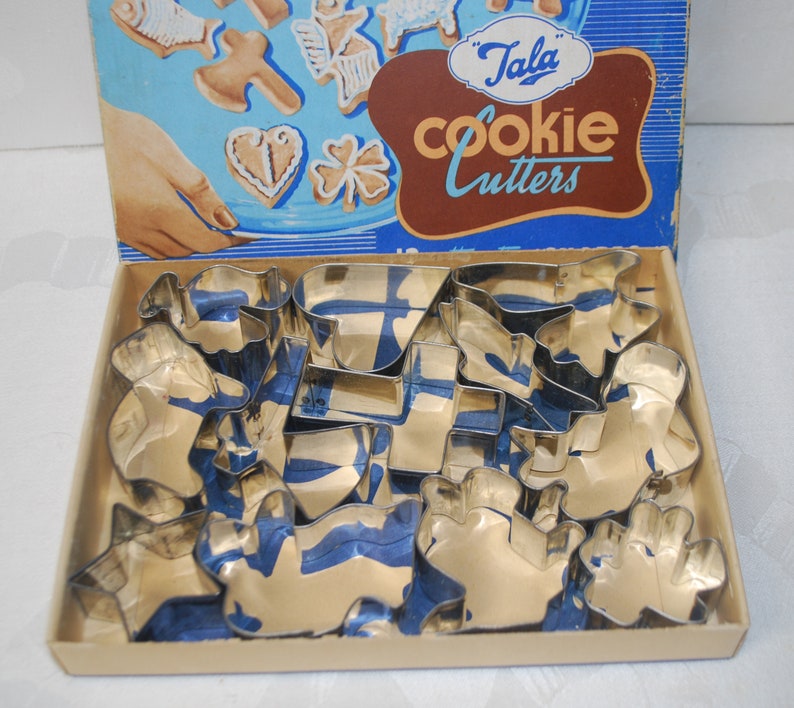 Puede incluir: Una caja vintage de cortadores de galletas Tala en una caja azul y blanca con el texto "Tala Cookie Cutters". La caja est&aacute; abierta y los cortadores de galletas est&aacute;n dispuestos en el interior. Los cortadores de galletas son de metal y tienen varias formas, incluyendo estrellas, corazones y animales.