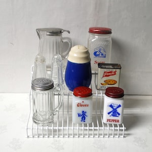 May include: A collection of vintage kitchenware, including glass salt and pepper shakers, a sugar jar, a blue and white shaker, and a small tin of French's Apple Pie Spice. The items are arranged on a white wire shelf.