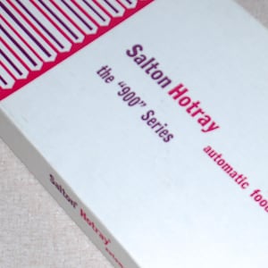 May include: A white rectangular box for a Salton Hotray automatic food warmer, 900 series. The packaging features purple text and a repeating geometric pattern in purple and white along the top edge, with the brand name printed on the side.