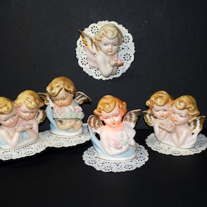 May include: Five porcelain cherub figurines with gold accents. The cherubs are wearing white and blue robes and have blonde hair. Some of the cherubs are holding flowers. The figurines are displayed on white doilies.