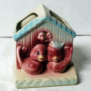 May include: A vintage ceramic birdhouse planter in shades of red, blue, and cream. The planter features two red birds with blue eyes and a small bird on top, all under a house-shaped roof. The base is a light cream color.