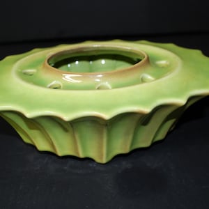 May include: A green ceramic flower frog with a scalloped edge and a central well with multiple holes. The frog has a light green glaze with a subtle brown tint.