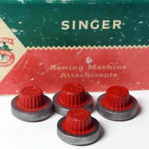 May include: Four red sewing machine attachments with a silver base are in front of a vintage box. The box is green and red with the word "SINGER" and "Sewing Machine Attachments" printed on it. The attachments have a ribbed design.