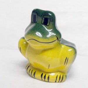 May include: A ceramic frog figurine with green and yellow glaze. The frog has black eyes and a black mouth.