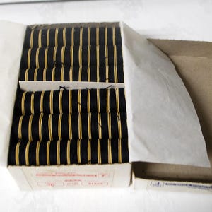 May include: An open cardboard box filled with multiple spools of black thread with gold-colored centers. The spools are neatly arranged in rows. The box is partially covered with white paper.