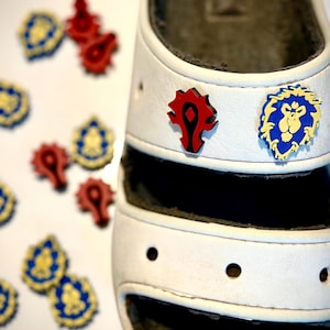 Op de afbeelding: Witte sandalen met een rood en blauw World of Warcraft-logo op de band. Er zijn verschillende andere World of Warcraft-logo's verspreid rond de sandalen.
