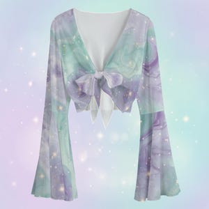 Op de afbeelding: Een crop top met lange, uitlopende mouwen en een V-hals, vastgemaakt met een strik. De stof toont een wervelend galaxypatroon in paars, teal en wit. Het ontwerp heeft een transparante kwaliteit, waardoor een etherische esthetiek ontstaat.