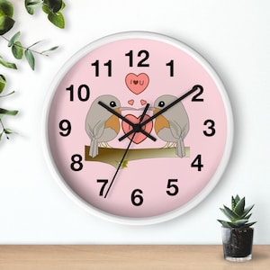 Könnte beinhalten: Rosa runde Wanduhr mit weißen Zahlen und einem weißen Rahmen. Zwei Cartoon-Vögel sitzen auf einem Ast und teilen sich ein Herz, auf dem "Ich liebe dich" steht.