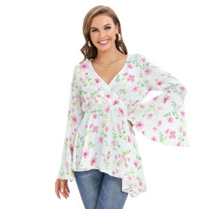 May include: A white wrap top with a V-neck and long, flared sleeves. The top is adorned with a delicate floral pattern in pink and green. The model is wearing blue jeans.