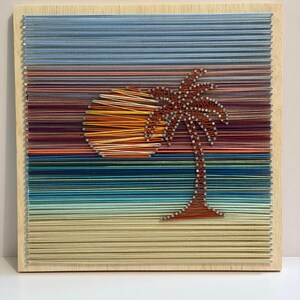 May include: String art wall decor featuring a sunset scene with a palm tree. The artwork uses colorful threads to depict the sun, sky, and beach. The palm tree is brown, and the sun is orange and yellow.