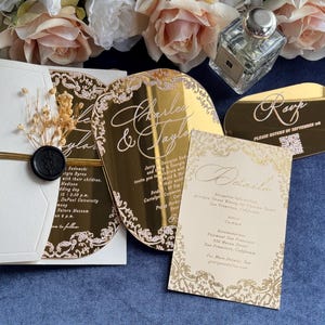 May include: A luxury wedding invitation suite in gold and white. The set includes a gold acrylic invitation, RSVP card, and details card. Floral embellishments and a wax seal add to the sophisticated design, ideal for a special occasion.