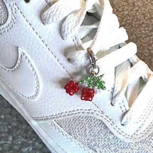 May include: A white trainer with white laces. A beaded cherry charm with red cherries and green leaves hangs from the laces. The trainer has a white leather upper and a white sole.
