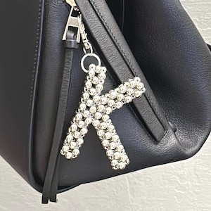 Puede incluir: Bolso de mano de cuero negro con cremallera plateada y un charm decorativo con la letra "K" plateada. El charm está hecho de pequeñas cuentas redondas plateadas. El bolso tiene un diseño simple y elegante.