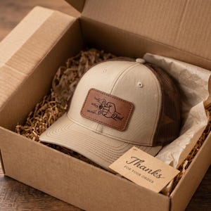 Op de afbeelding: Een beige en bruine baseballcap met een leren patch met de tekst "Papa" en de namen "Emily", "Willow" en "Michael". De cap zit in een kartonnen doos met een kaartje "Bedankt voor uw bestelling".