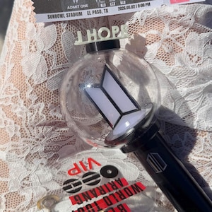 May include: A black and clear light stick with the word "J-HOPE" on top. The light stick has a white and black logo and is next to a ticket that says "Sunbowl Stadium El Paso, TX". A VIP pass is also visible.