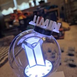 May include: A clear, spherical light-up device with a white BTS logo and the name "JIMIN" on top. The device emits a soft, blue glow. The base is black, and the overall design is modern and sleek.