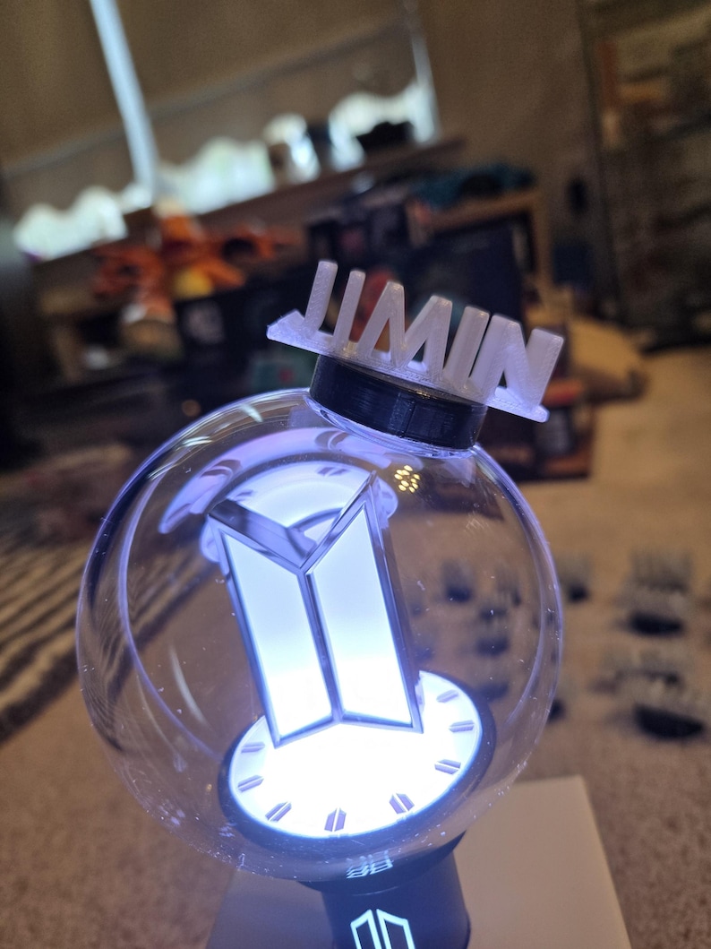 May include: A clear, spherical light-up device with a white BTS logo and the name "JIMIN" on top. The device emits a soft, blue glow. The base is black, and the overall design is modern and sleek.