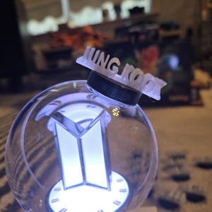 May include: A clear, spherical light-up device with a white and silver logo and the name "JUNG KOOK" on top. The device emits a soft, white glow, and the base is black.