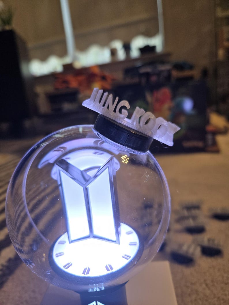 May include: A clear, spherical light-up device with a white and silver logo and the name "JUNG KOOK" on top. The device emits a soft, white glow, and the base is black.