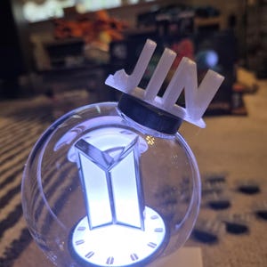 May include: A clear, spherical light-up device with a white BTS logo and the name "JIN" on top. The device has a black base and emits a soft, white glow. The overall design is modern and sleek.