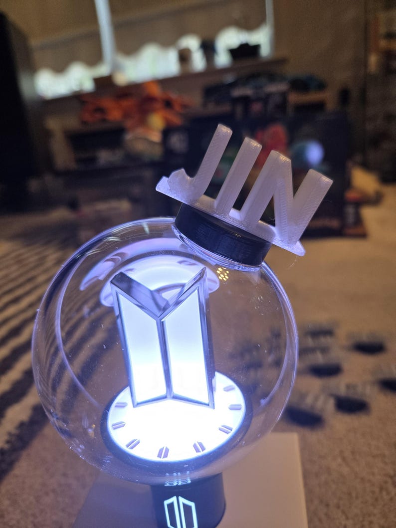May include: A clear, spherical light-up device with a white BTS logo and the name "JIN" on top. The device has a black base and emits a soft, white glow. The overall design is modern and sleek.