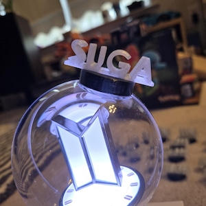 May include: A clear, spherical light stick with a white, illuminated logo and the name "SUGA" on top. The light stick has a black base and a white, glowing interior, creating a bright, eye-catching effect.