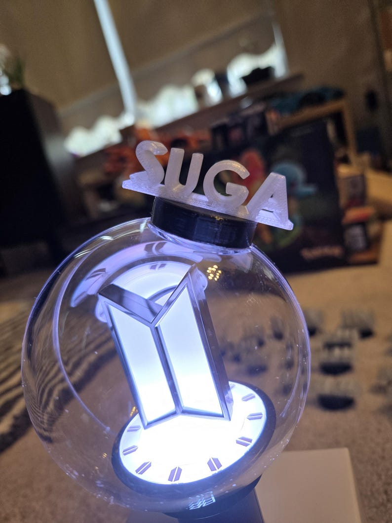 May include: A clear, spherical light stick with a white, illuminated logo and the name "SUGA" on top. The light stick has a black base and a white, glowing interior, creating a bright, eye-catching effect.