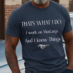 May include: Navy blue t-shirt with the text "THAT'S WHAT I DO: I work on Mustangs. And I know Things" and a Mustang logo. The person is wearing a brown cap and sunglasses, leaning against a classic blue car.