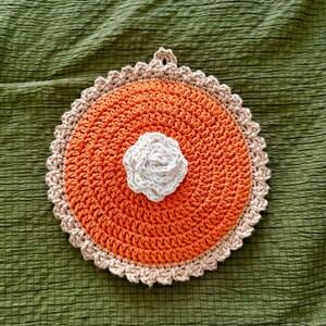 Peut inclure: Un dessous de plat fait à la main en crochet en forme de tarte. Le dessous de plat présente un centre orange, une croûte beige et une fleur blanche au centre. Il y a une boucle pour l'accrocher. Le fond est un tissu vert texturé.