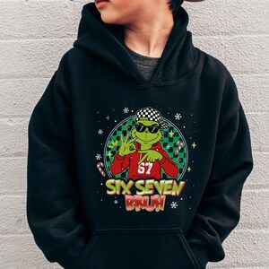 May include: Black hoodie featuring a cartoon frog wearing sunglasses and a backwards checkered hat, giving a peace sign. The frog is wearing a red shirt with the number 67. The text "SIX SEVEN BRUH" is below the frog.