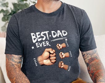 May include: Dark gray t-shirt with the text "BEST DAD EVER" in white. The shirt features illustrations of fists and small arms with names. The names are Ethan, Kevin, Emma, and Noah. The shirt is made of a soft material.