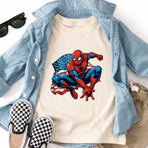 May include: A cream-colored t-shirt featuring a graphic of Spider-Man with an American flag. The shirt is paired with a light blue denim shirt, khaki shorts, black sunglasses, and black and white checkered slip-on shoes.
