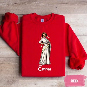 May include: A bright red sweatshirt featuring a graphic of a woman in a white gown with a yellow sash. The name "Emma" is written in white script below the graphic. The word "RED" is in a pink rectangle in the lower right corner.