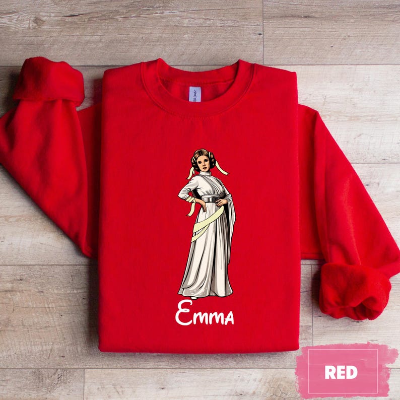 May include: A bright red sweatshirt featuring a graphic of a woman in a white gown with a yellow sash. The name "Emma" is written in white script below the graphic. The word "RED" is in a pink rectangle in the lower right corner.
