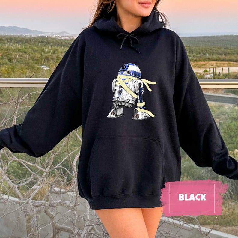 May include: Black hooded sweatshirt featuring a graphic of the Star Wars character R2-D2 wrapped in yellow bandages. The design is centered on the front of the sweatshirt, which has a kangaroo pocket and a drawstring hood. The word "BLACK" is in a pink rectangle.