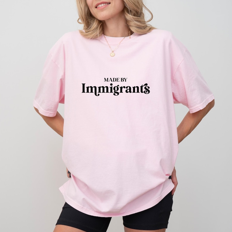 Może przedstawiać: R&oacute;żowy T-shirt z napisem "MADE BY Immigrants" w kolorze czarnym.