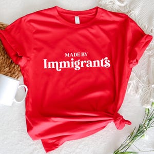 Może przedstawiać: Czerwona koszulka z białym napisem "Made by Immigrants".