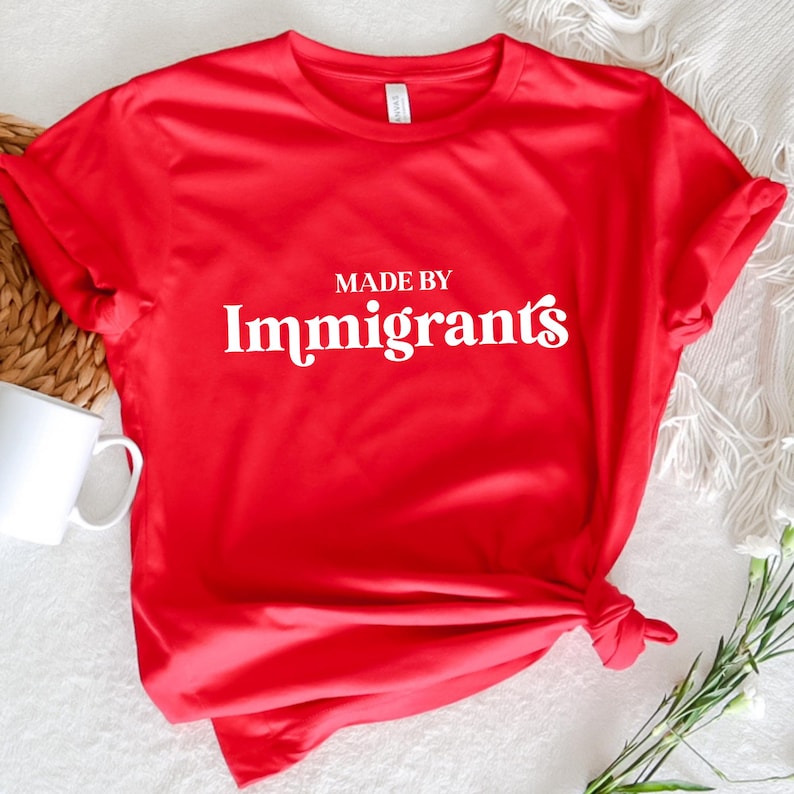 Może przedstawiać: Czerwona koszulka z białym napisem "Made by Immigrants".