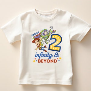 Puede incluir: Camiseta blanca de manga corta con un gr&aacute;fico colorido de Woody y Buzz Lightyear de Toy Story. El gr&aacute;fico incluye el n&uacute;mero "2", las palabras "infinity & BEYOND" y el nombre "DARIAN".