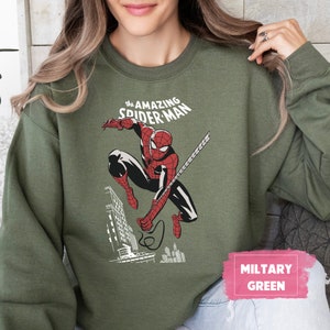 Pu&ograve; includere: Una felpa verde con una grafica di Spider-Man che si dondola attraverso la citt&agrave;. Il testo "The Amazing Spider-Man" &egrave; stampato sopra la grafica. La felpa &egrave; etichettata "Verde Militare".