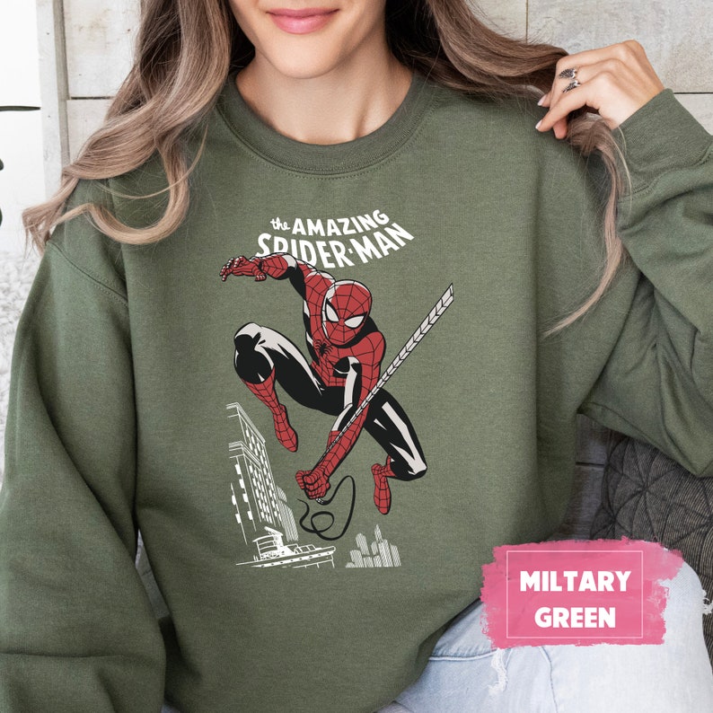 Pu&ograve; includere: Una felpa verde con una grafica di Spider-Man che si dondola attraverso la citt&agrave;. Il testo "The Amazing Spider-Man" &egrave; stampato sopra la grafica. La felpa &egrave; etichettata "Verde Militare".