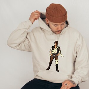 May include: A beige hoodie featuring a graphic of a character from a science fiction film. The character is wearing a brown vest, pants, and boots, with a white sash and a holster. The person in the image is wearing a brown beanie.